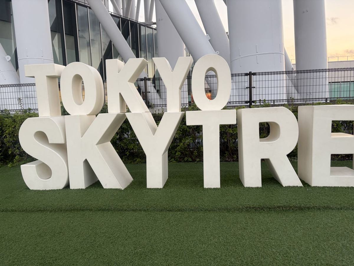 人工芝の上に並ぶ白い立体文字のモニュメント「TOKYO SKYTREE」。背後にスカイツリーの白い脚が見える